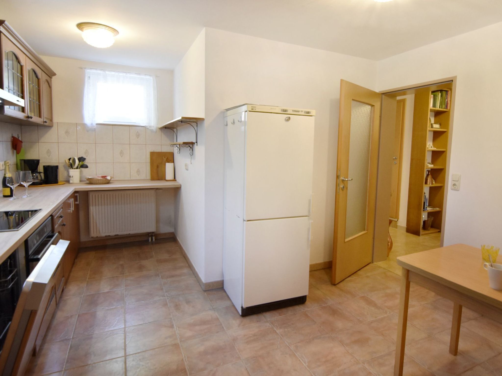 A compact kitchen with a refrigerator, a window, a radiator, and a wooden door leading to a pantry.