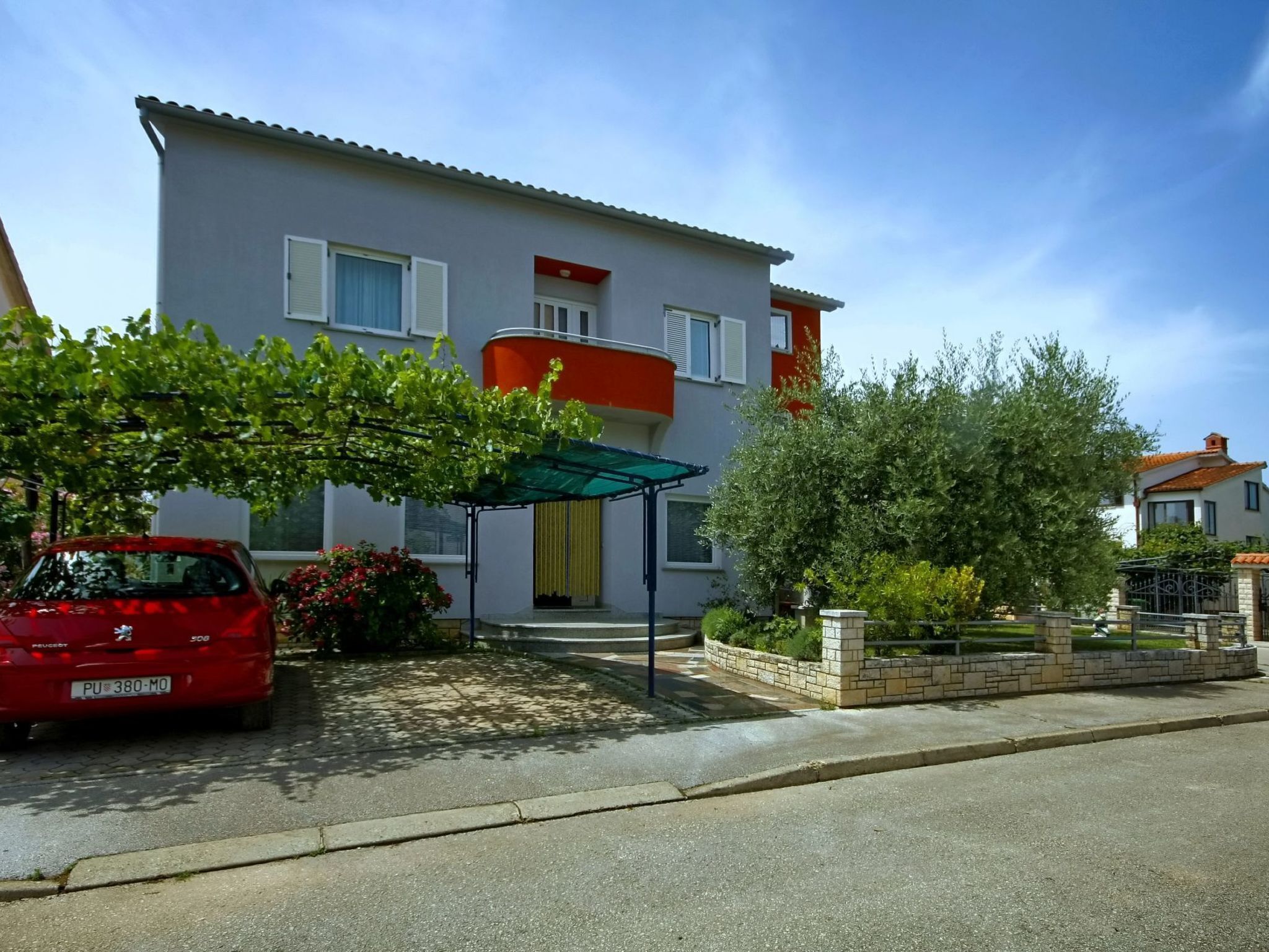 A two-story house with a covered porch, a parked red car, and a garden with various plants and a small tree.