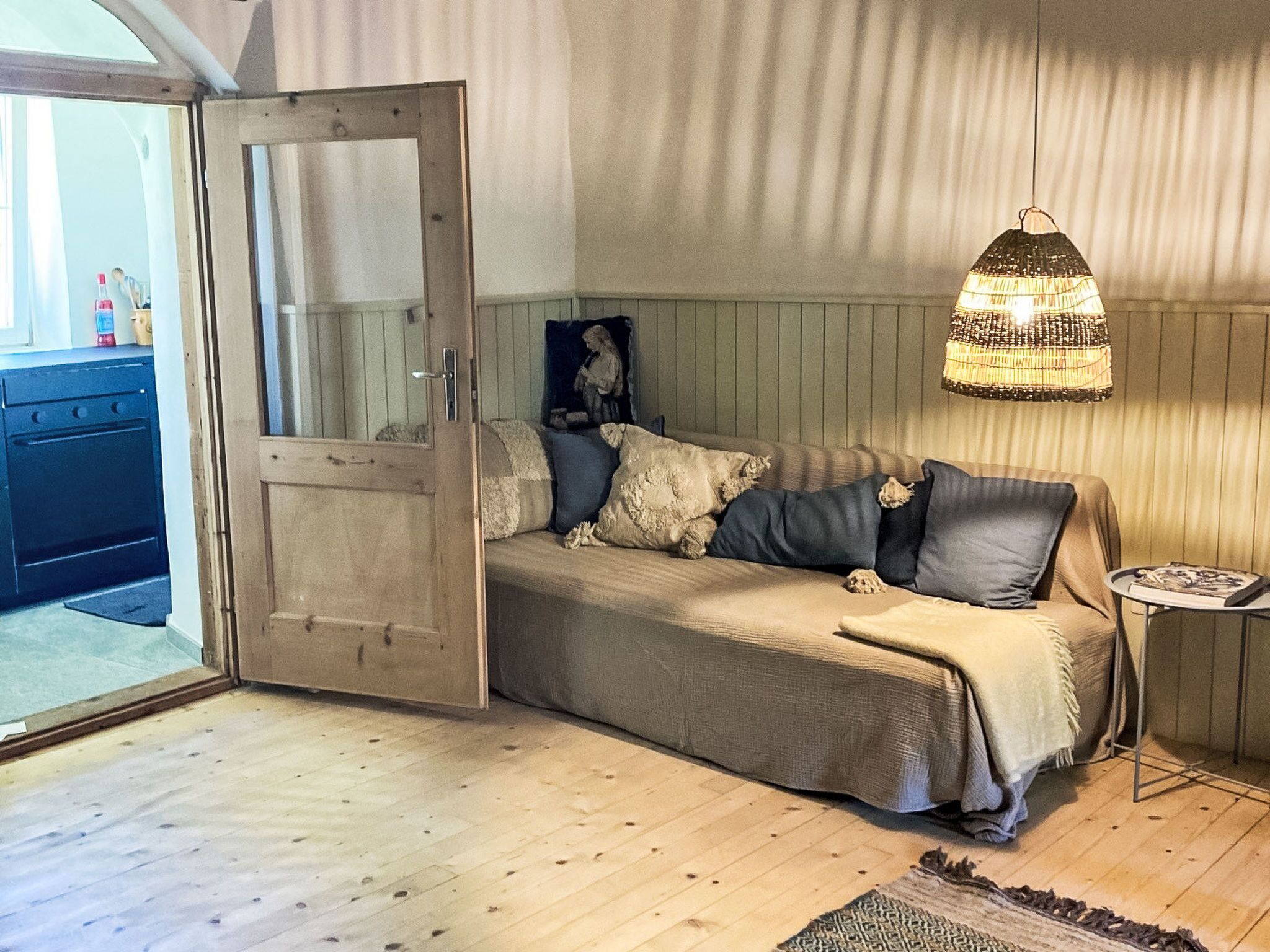Une pièce chaleureuse avec un plancher en bois, un lit recouvert d’une couverture beige, une lampe fixée au mur et une petite table sur laquelle repose un magazine.