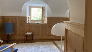 A room with wooden paneling, a white radiator, a small table with a book, a blue lamp, and a window with a view of greenery. - Chesa alb sco'l floc by Interhome (S-chanf)