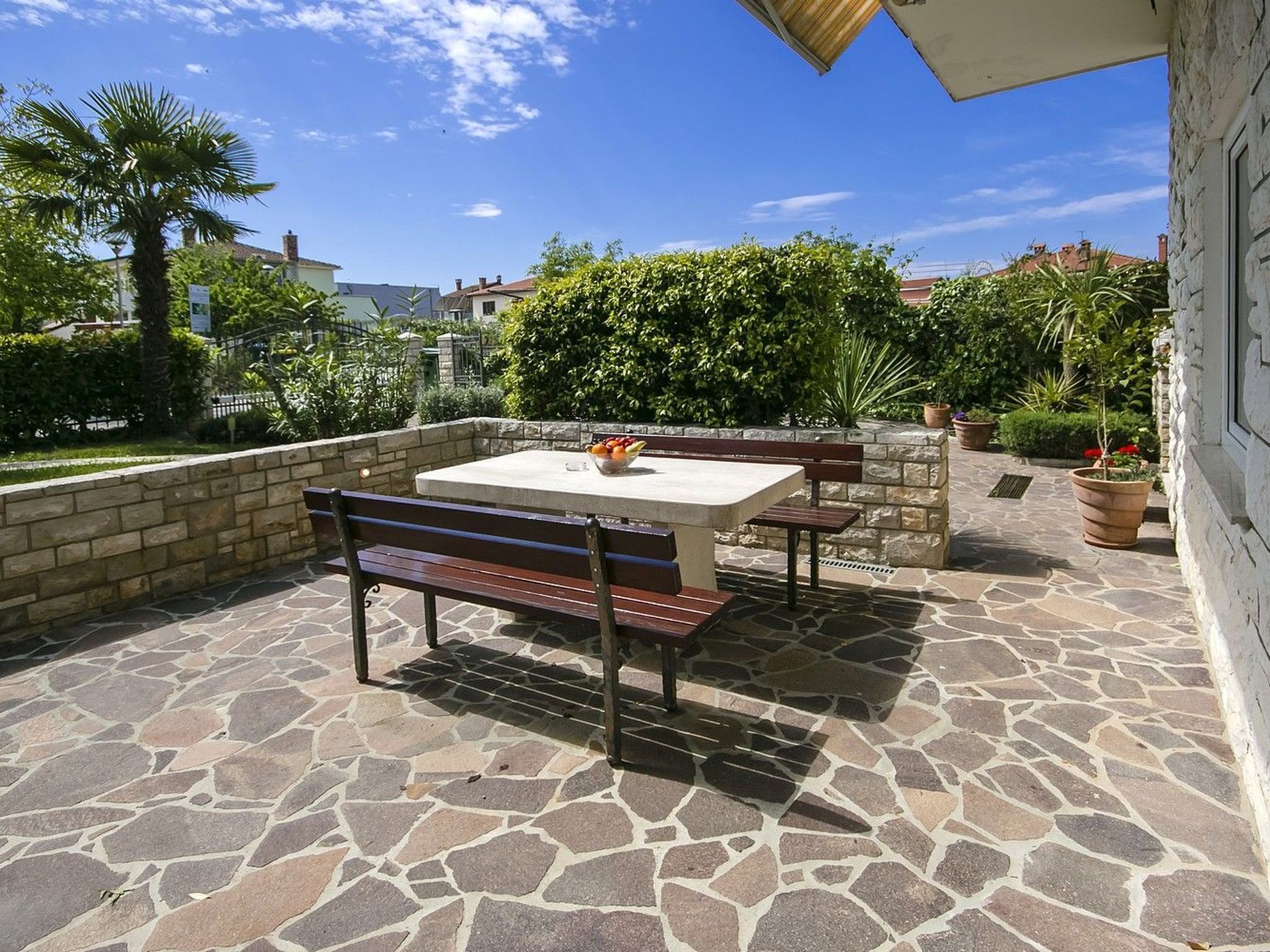 An outdoor patio with stone flooring, a wooden bench, a table with fruit, and potted plants.