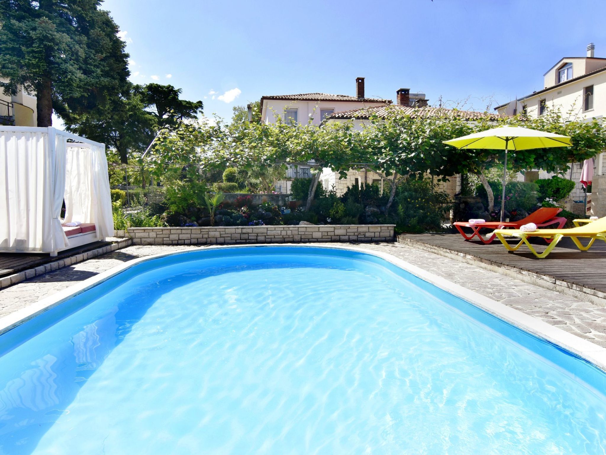 A swimming pool with lounge chairs and an umbrella, surrounded by greenery and buildings.