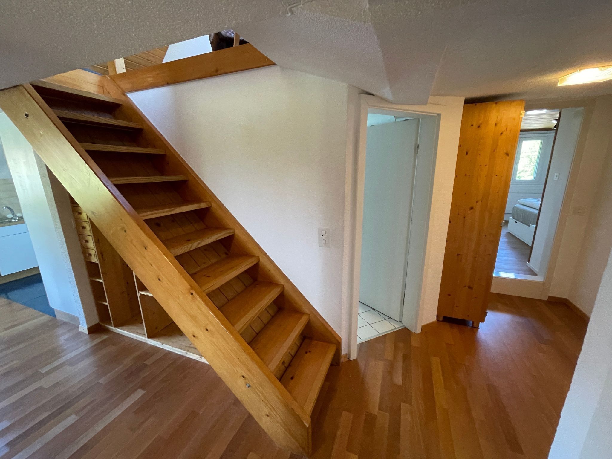 Un escalier en bois menant à un deuxième étage, un couloir avec une porte, et une vue sur une autre pièce où se trouve un lit.