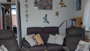 En stue med en grå sofa, to lænestole, et glasbord og dekorative billeder på væggen, blandt andet sommerfugle og et gruppesbillede.