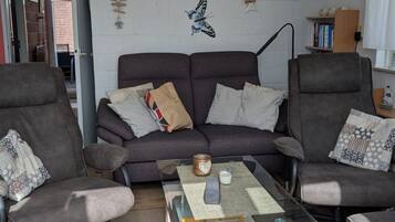 Un salon avec un canapé gris, deux fauteuils, une table basse en verre et des œuvres d’art décoratives murales, notamment des motifs de papillons et une scène de foule.