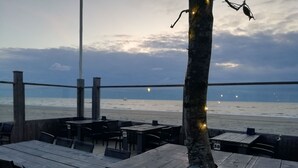 En udendørs siddeplads med træborde og stole, med udsigt over en strand og en sejlbåd i horisonten.