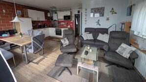 En moderne stue med sofa, lænestole og sofabord. I baggrunden er der et køkken med mikroovn og køleskab.