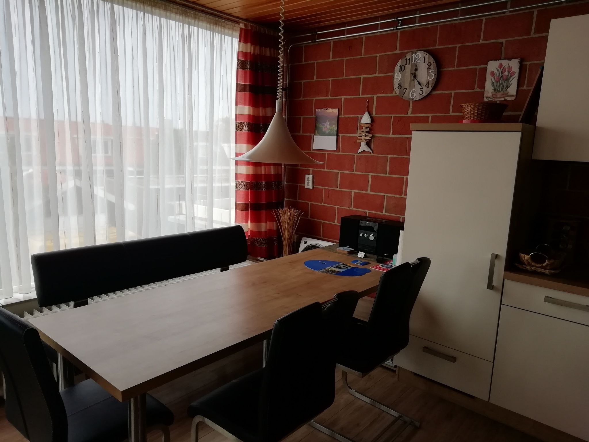 Une cuisine avec une table en bois, des chaises noires, un luminaire suspendu, une horloge et un mur en briques.