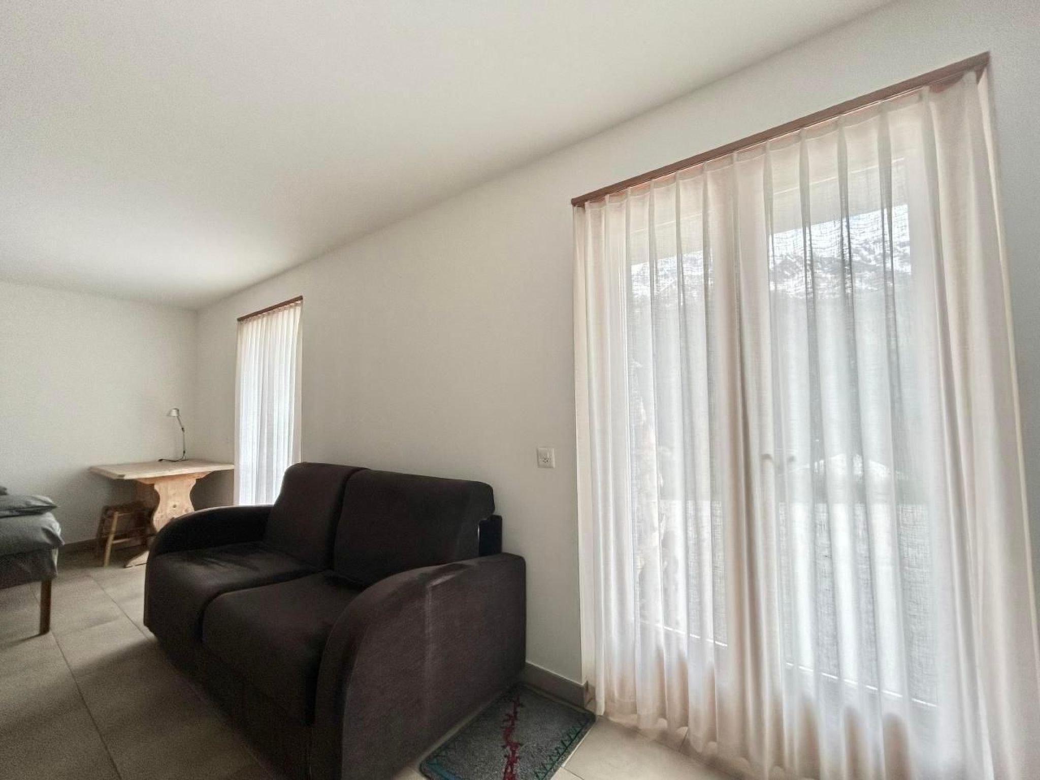 A living room with a sofa, a small table, and a chair. There are large windows with sheer curtains.