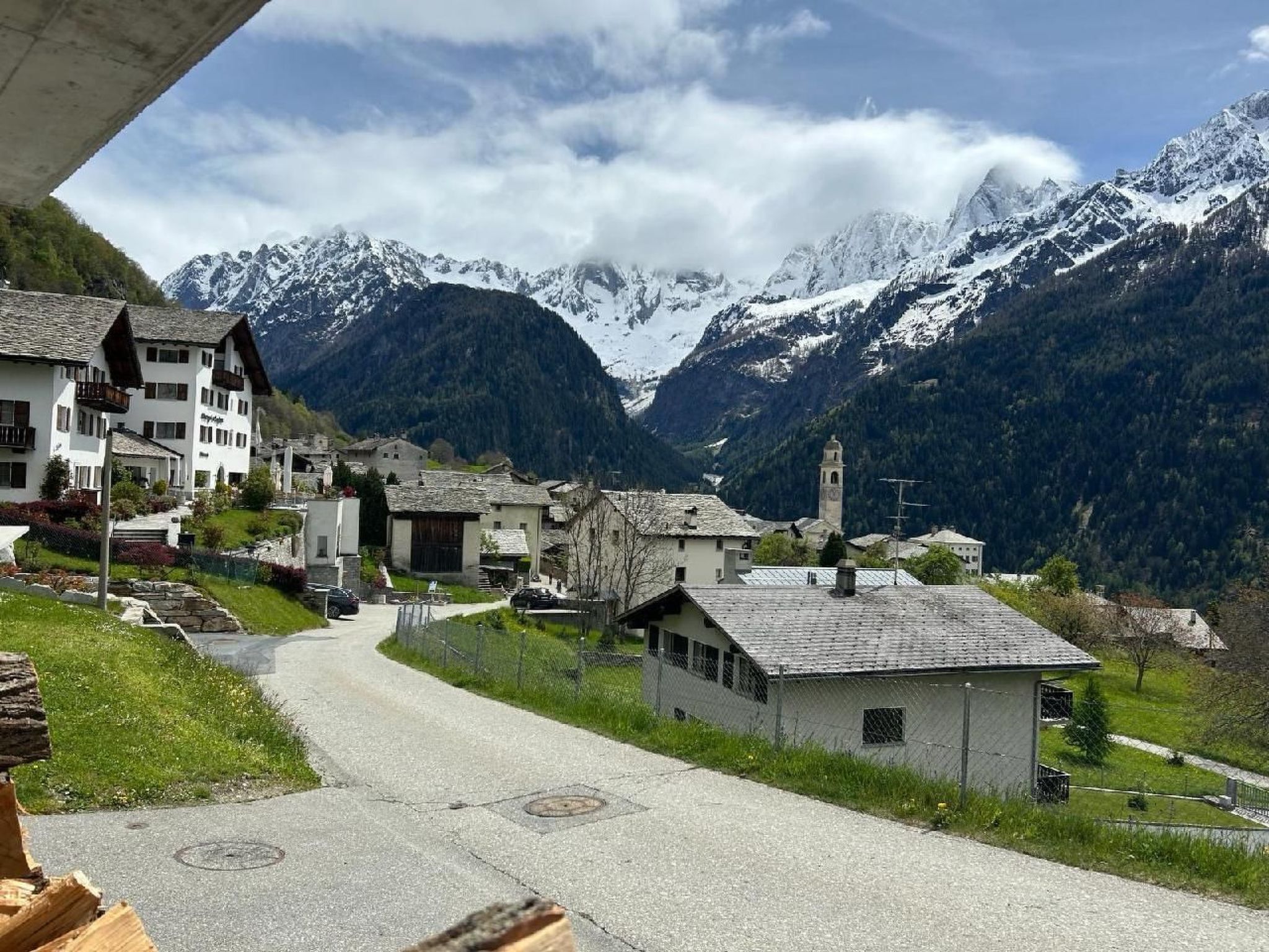 A mountain village with traditional buildings, a paved road, and snow-capped mountains in the background.