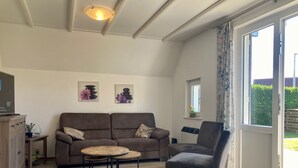 Een woonkamer met een bank, fauteuil en een ronde tafel. Er is een televisie op een kast, kunst aan de muur en een raam met gordijnen.