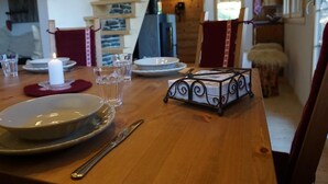 Une table à manger en bois est dressée avec des assiettes, des verres et des couverts. Une bougie est allumée sur la table, et un plateau en métal contenant des journaux y est posé.