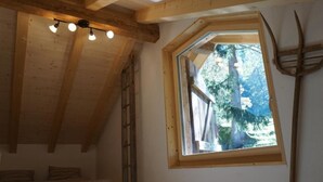 Une petite pièce rustique avec un plafond en pente, un lit en mezzanine, une échelle, une fenêtre donnant sur les arbres et une fourche accrochée au mur.