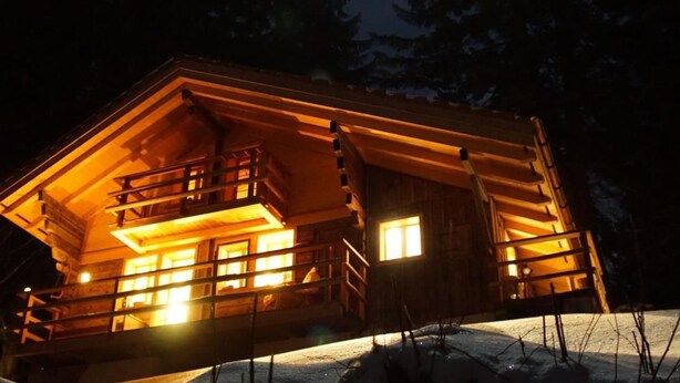 Une cabane en bois dotée d’un balcon, de fenêtres illuminées et entourée de neige.