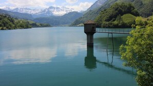 Un lago tranquilo con una pequeña torre sobre pilotes en el centro, rodeado de colinas verdes exuberantes y montañas nevadas a lo lejos.