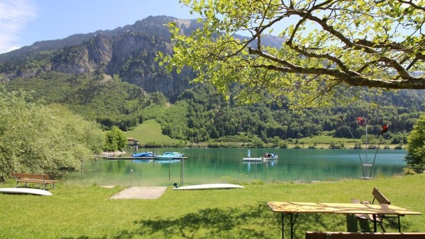 Un área de picnic junto al lago con bancos y una mesa, barcos en el agua y montañas al fondo.