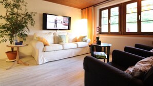 Una sala de estar con un sofá blanco, un sillón oscuro, un televisor en la pared, una planta en maceta y una ventana con cortinas.