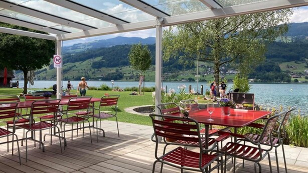 An outdoor patio with red metal chairs and tables, overlooking a lake with mountains in the background. - Ferienhaus Flüeli by Interhome (Flüeli-Ranft)