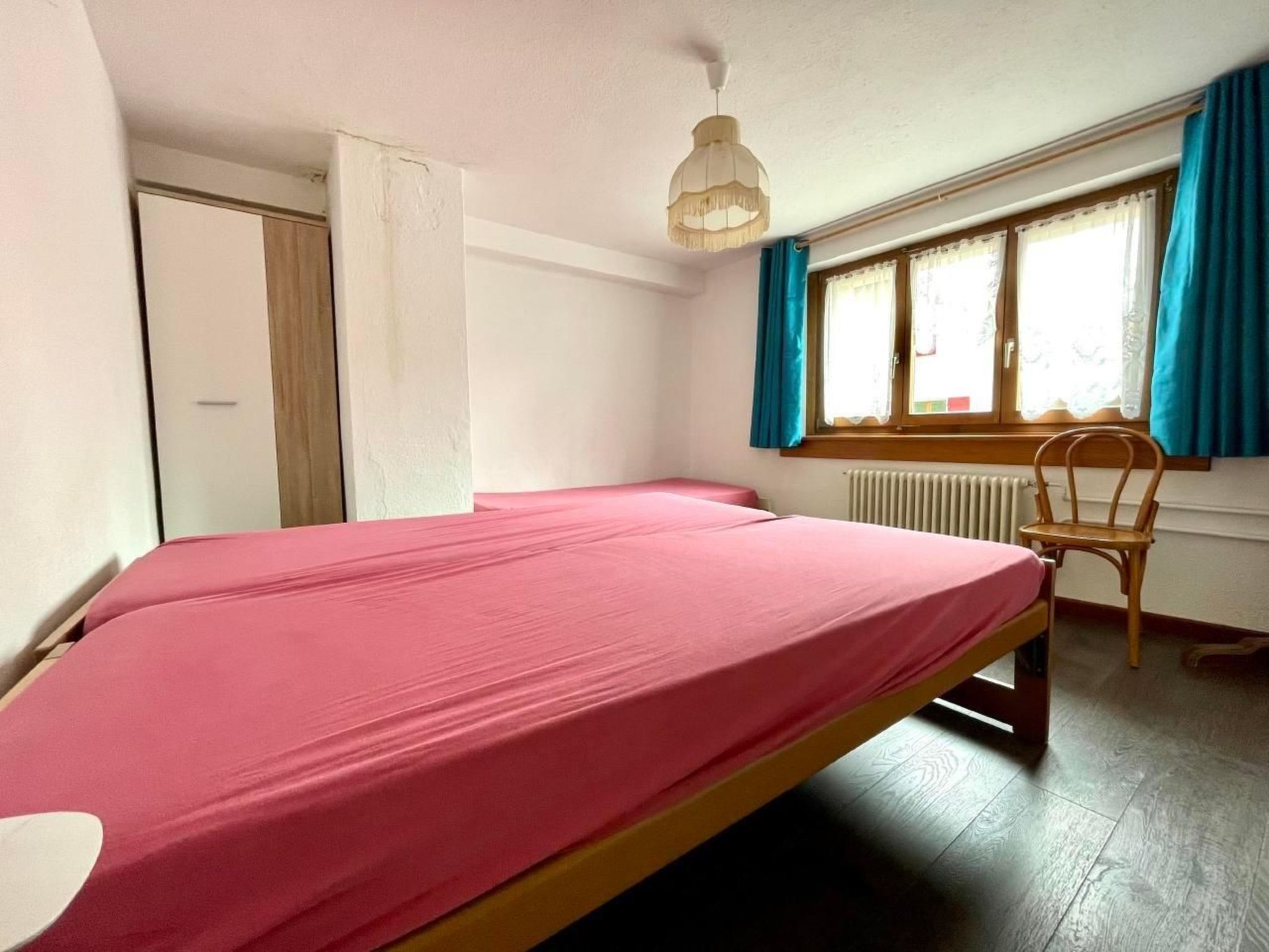 A single bed with a pink mattress in a room with a wooden floor, a window with blue curtains, and a radiator.