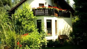 Ein zweistöckiges Haus mit Balkon, umgeben von üppigem Grün und roten Blüten.