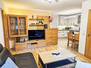 A modern living room with a flat-screen TV, wooden cabinets, a dining area with a table and chairs, and a kitchen with white cabinets and appliances. - Fewo Ginkgo by Interhome (Dresden)