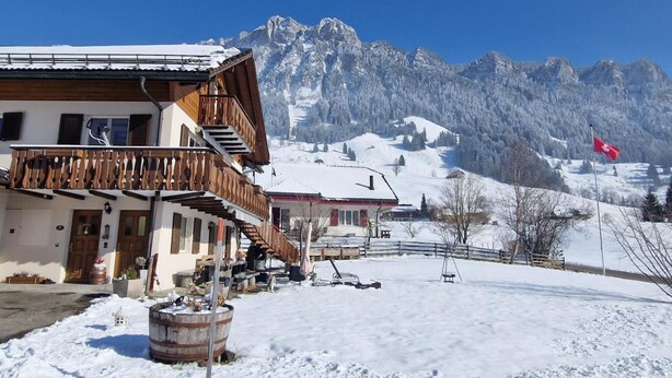 Un bâtiment de style chalet, doté d’un balcon en bois, avec une montagne enneigée en arrière-plan et un drapeau suisse.