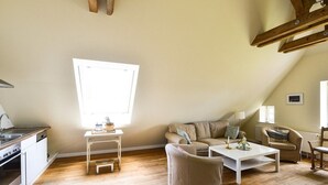 Ein gemütliches Loft mit Wohnbereich und Küchenzeile, Holzbalken, einem großen Fenster und einem weißen Couchtisch.