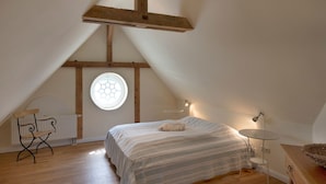 Ein Schlafzimmer mit schrägen Wänden, einem Bett mit gestreifter Bettdecke, einem Rundfenster, einem kleinen Tisch und einem Stuhl.