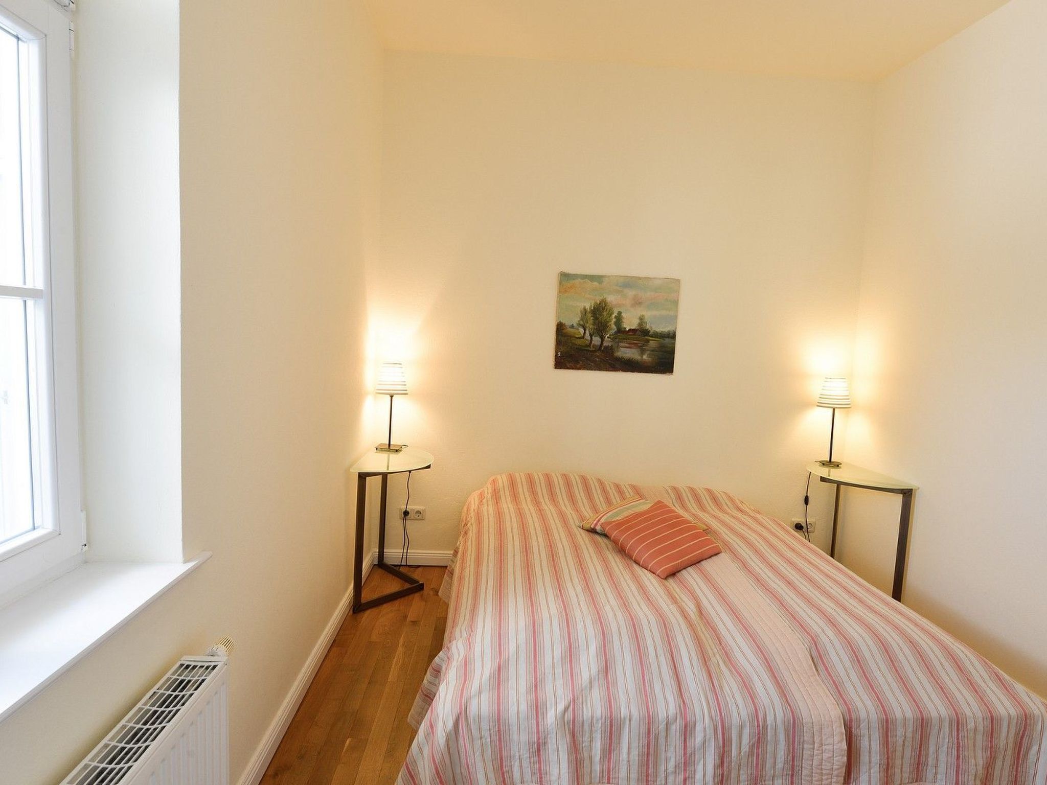 Une petite chambre bien éclairée, avec un lit simple, deux lampes de chevet, un radiateur et une fenêtre.
