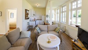 A spacious living area with a sofa, a coffee table, and a TV. There is a kitchen in the background with white cabinets and a dining table. - Wohnung im ersten Obergeschoss des Gutshauses by Interhome (Gingst)