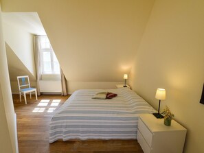 A bedroom with a sloped ceiling, a bed with striped bedding, a chair, bedside tables with lamps, and a window with curtains. - Wohnung im zweiten Obergeschoss des Gutshauses by Interhome (Gingst)