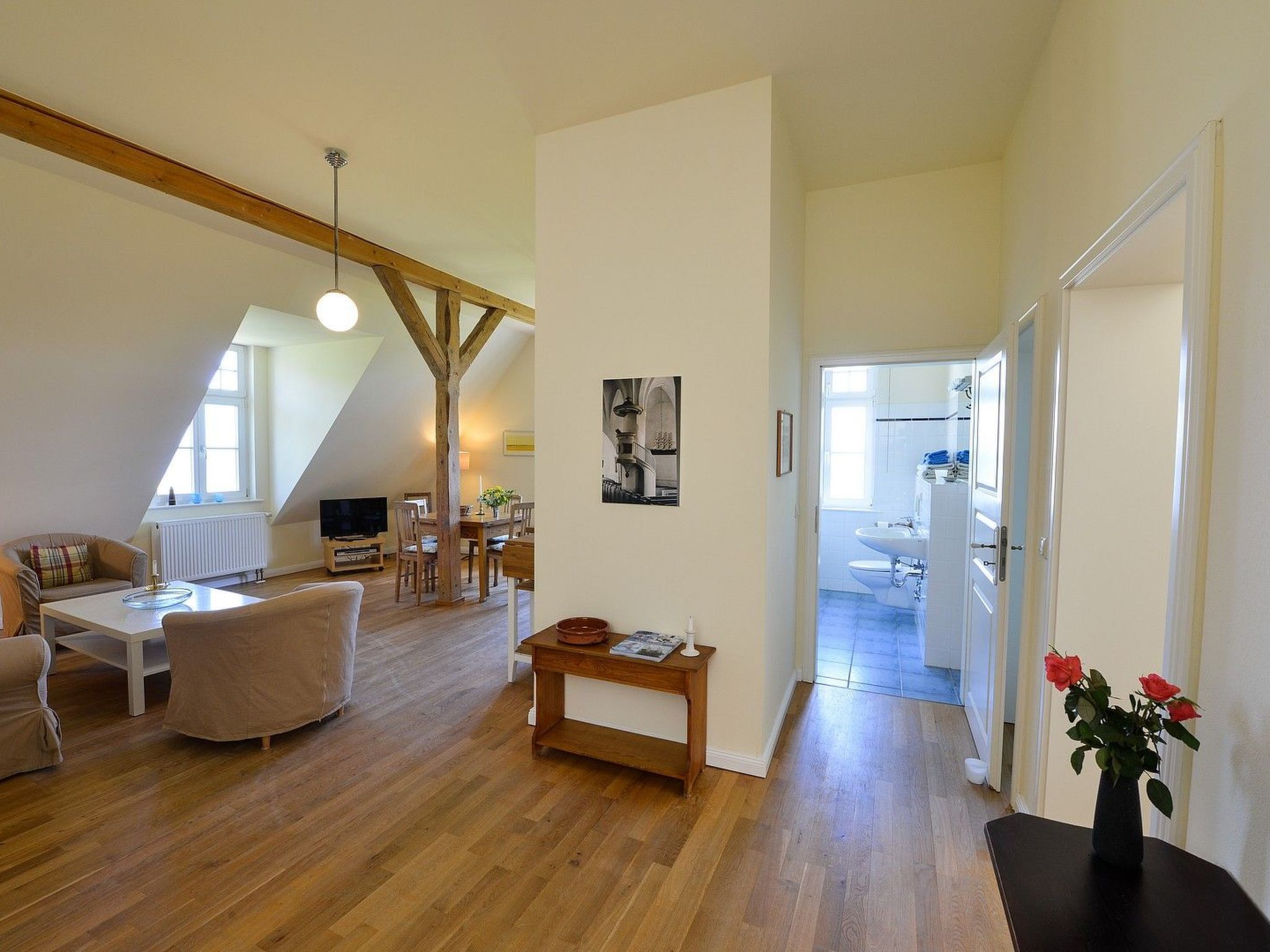 A spacious loft apartment with a high ceiling, exposed wooden beams, a dining area, a living room with a sofa and a TV, and a kitchen visible through an open door.
