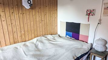 En enkeltseng med et hvidt tæppe, en trævæg, en lampe på væggen og et lille bord med en lampe.