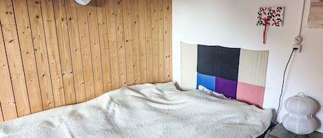 Ein Einzelbett mit weißer Decke, eine Holzwand, eine Wandleuchte und ein kleiner Tisch mit Lampe.