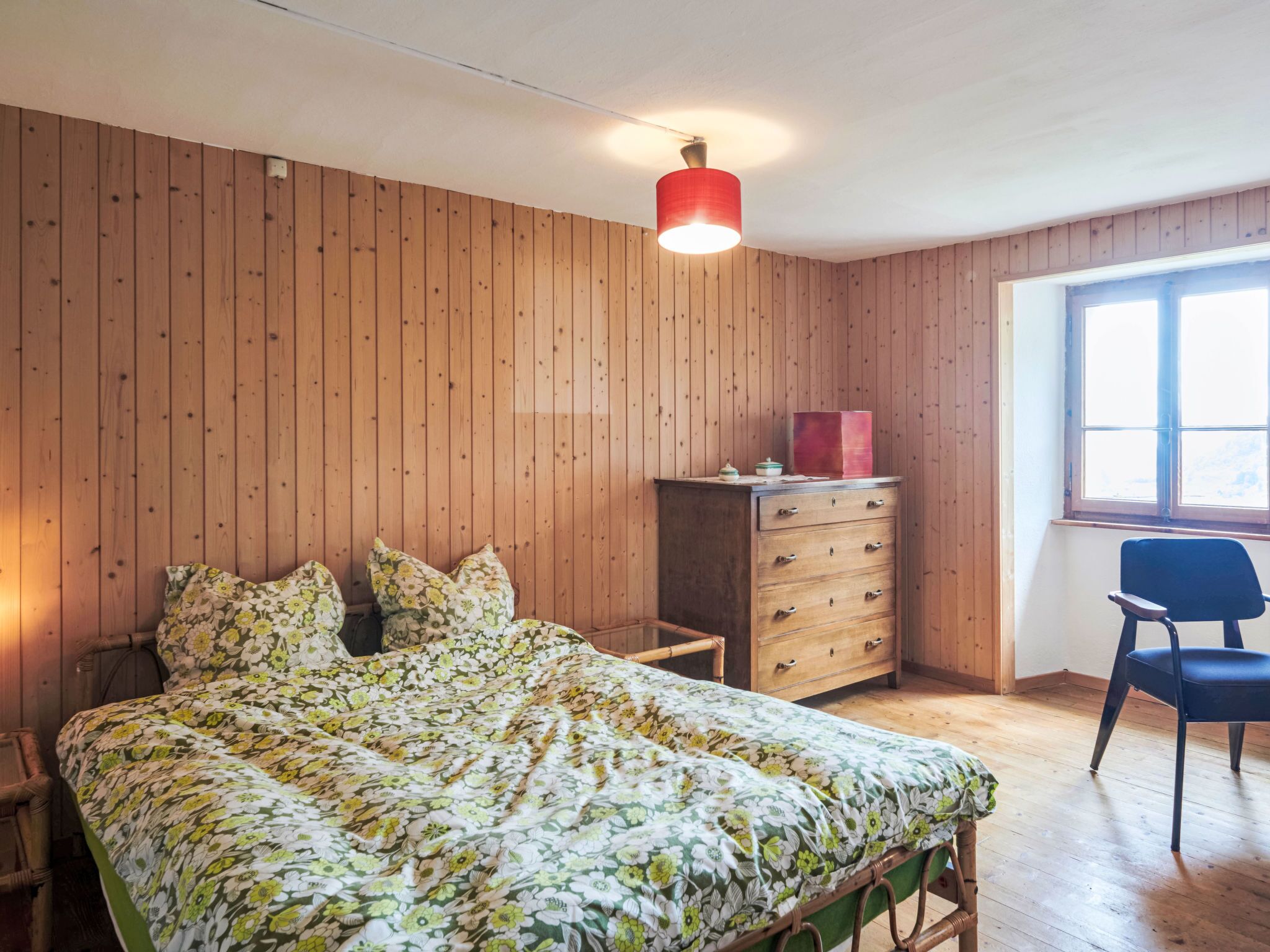 Une chambre avec un mur lambrissé en bois, un lit avec une literie à motifs floraux, une commode, une chaise et une fenêtre.