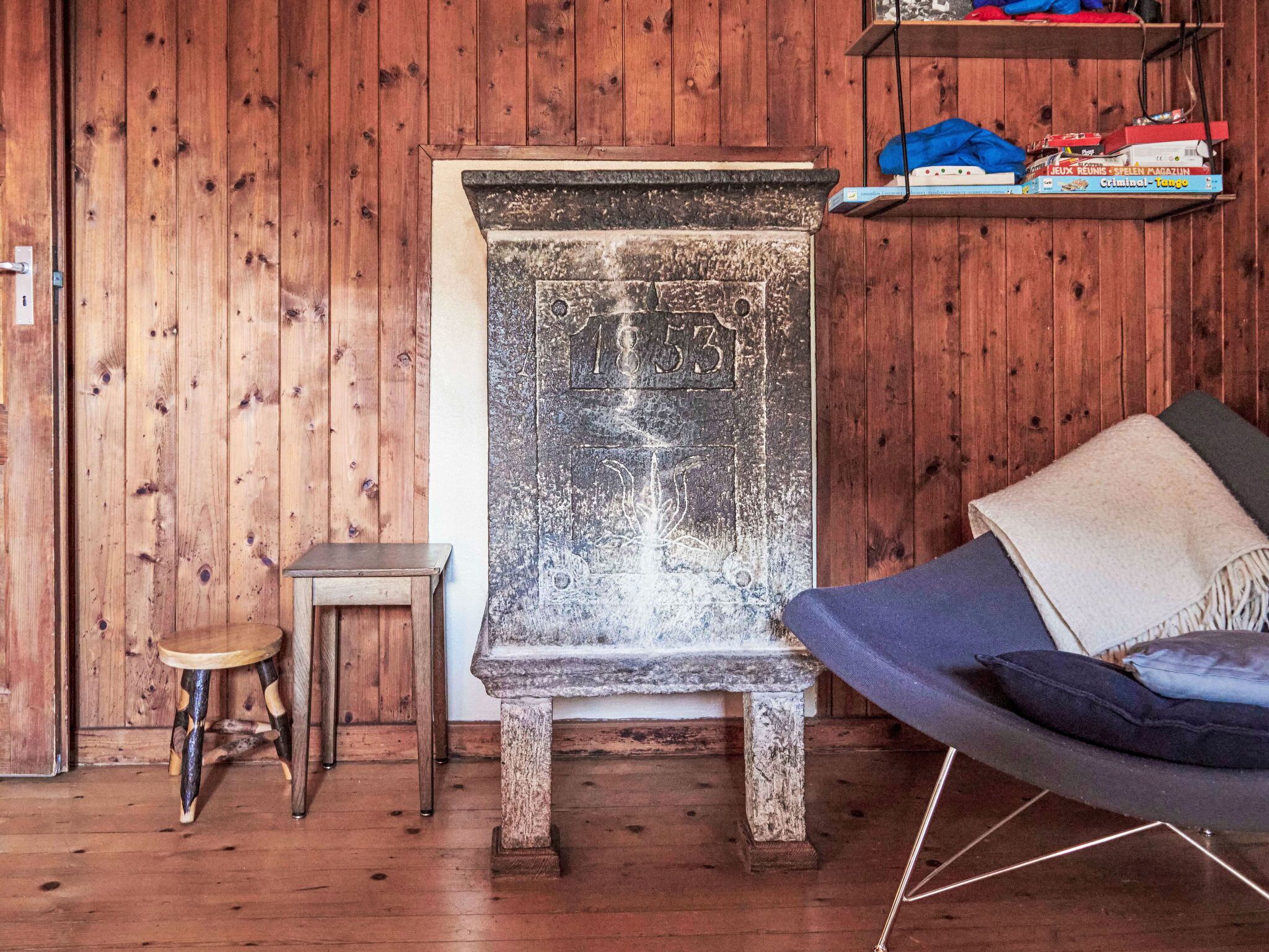 Une pièce rustique avec un plancher en bois, une cheminée en pierre ornée d’une plaque commémorative, un petit tabouret en bois, une chaise bleue avec des coussins et une étagère remplie de livres et de jouets.
