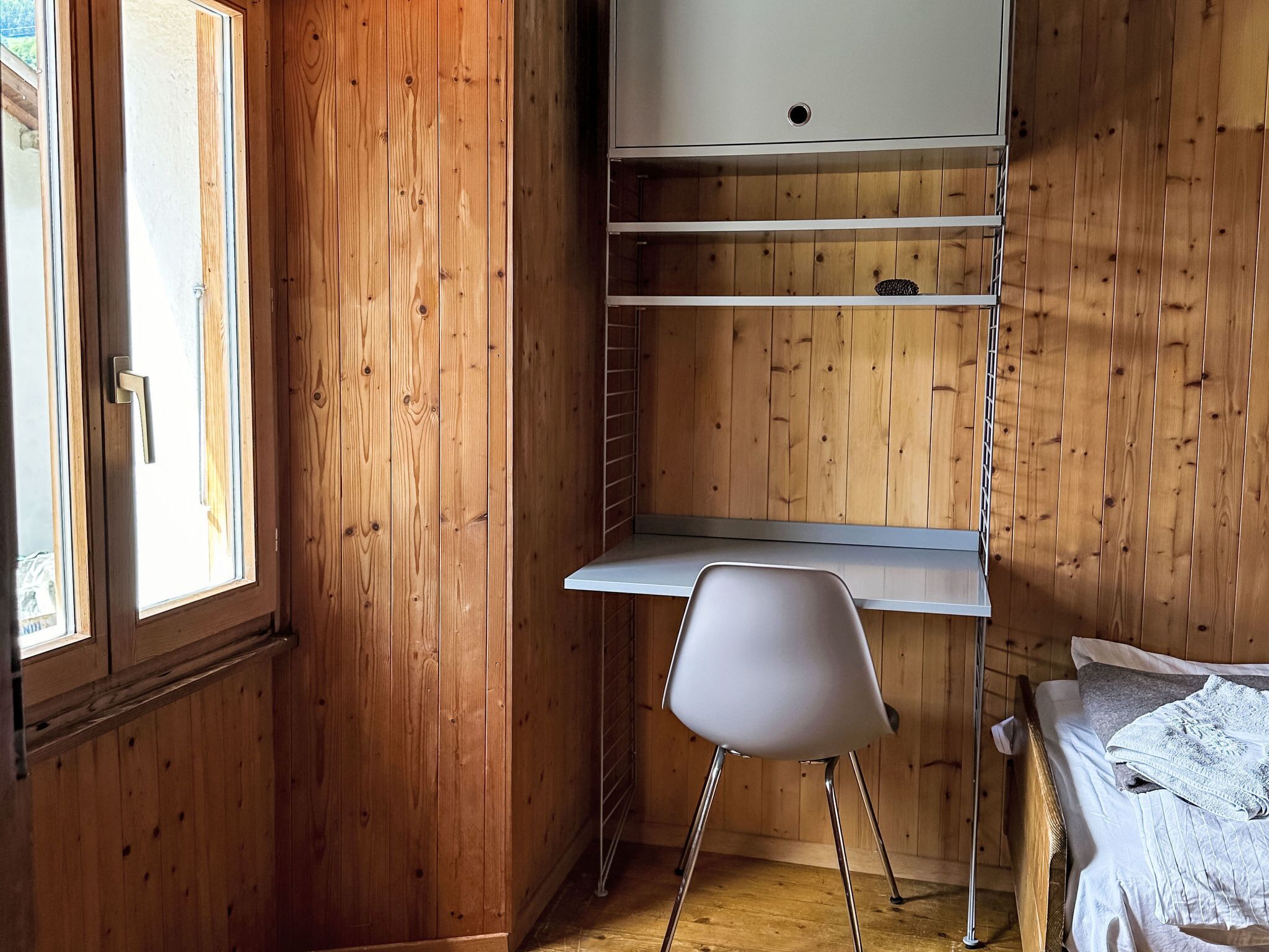 Una habitación con paredes de madera, un escritorio blanco con estantes, una silla blanca y una cama con manta blanca.