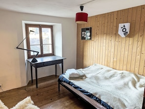 A bedroom with a wooden wall panel, a desk with a lamp, a bed with a blanket, and a window.