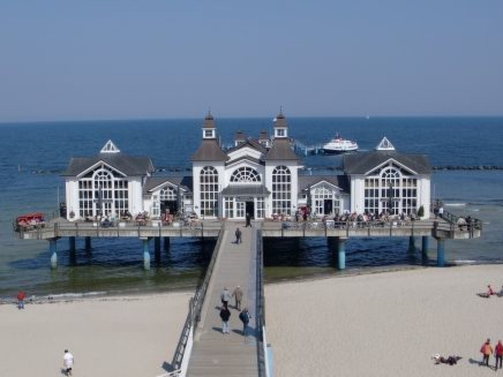 A pier with a white building featuring multiple windows and a central dome, extending over a sandy beach.
