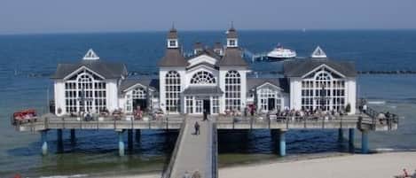 A pier with a white building featuring multiple windows and a central dome, extending over a sandy beach.