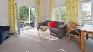 A spacious living room with a grey sofa, a white coffee table, and a dining table with wicker chairs. Large windows with yellow curtains provide ample natural light and views of greenery outside. - Villa Celia Sellin by Interhome (Ostseebad Sellin)