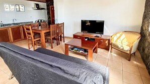 Een woonkamer met een bank, een televisie, een eettafel met stoelen en een keuken met een fornuis en kastjes.