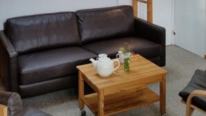 Una habitación acogedora con un sofá de cuero marrón, una mesa de centro de madera con una tetera blanca y un florero, un cuadro de un paisaje montañoso en la pared, y una escalera de madera apoyada contra una puerta blanca.