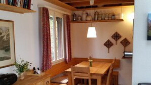 Un comedor acogedor con una mesa y sillas de madera, una planta pequeña en la mesa, y un estante con frascos y una lámpara.