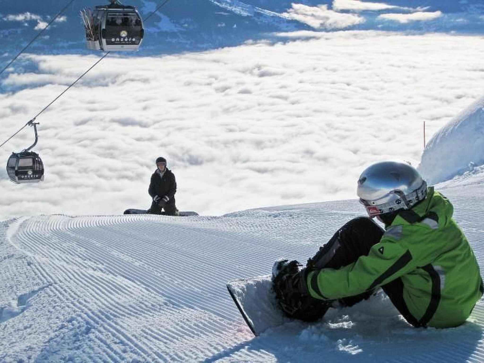 Un snowboarder sentado en una pendiente nevada con un telesilla al fondo.