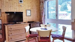 A cozy room with a wooden wall, a television, a small cabinet, a round table with chairs, and a large window with curtains. - Ski-in Wohnung in Anzere mit Parkplatz by Interhome (Anzère)