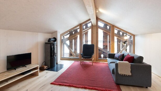 A modern loft bedroom with a large skylight, a cozy gray sofa, a red rug, and a TV on a wooden shelf. - Bijou - BioBergün by Interhome (Bergün)