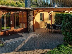 Eine überdachte Terrasse mit Steinboden, Holzmöbeln und Blick auf einen bewaldeten Hang.