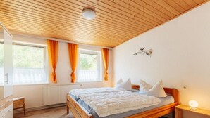 A bedroom with a wooden bed, a nightstand with a lamp, a dresser, and a window with curtains. - Vacation home Zita by Interhome (Thale)