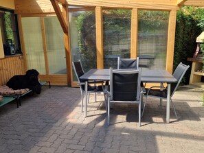 Eine Terrasse mit Tisch und Stühlen, ein Hund liegt auf einer Bank und ein Grill ist im Hintergrund zu sehen.